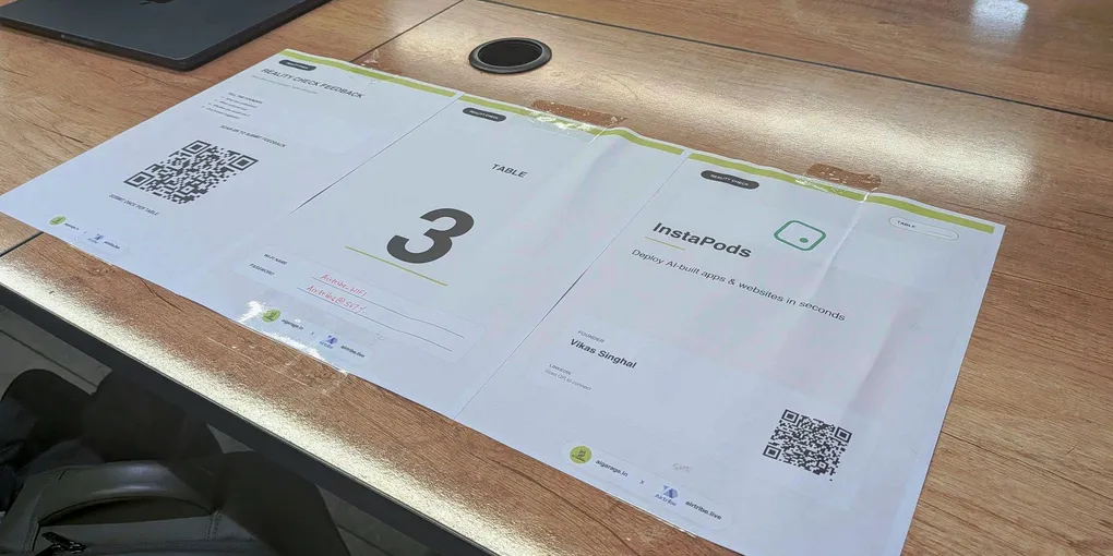 Three printed sheets taped down on a wooden meeting table: a Reality Check feedback form with a QR code, a card reading TABLE 3, and a placard reading InstaPods, Deploy AI-built apps and websites in seconds, founder Vikas Singhal. The wifi password is filled in by hand in red pen.